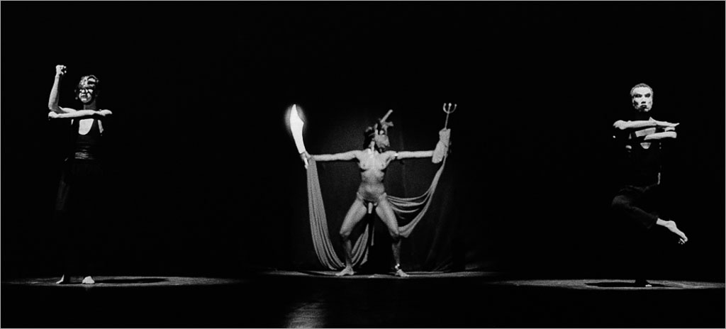 Uma performance dramática em preto e branco com três artistas. A artista central está usando uma máscara e um traje esvoaçante, estendendo os braços graciosamente parcialmente de pé. Os dois artistas laterais são pegos no meio do movimento, com braços e pernas equilibrados. O palco está com uma iluminação suave e focada nas figuras. Foto: Guto Muniz