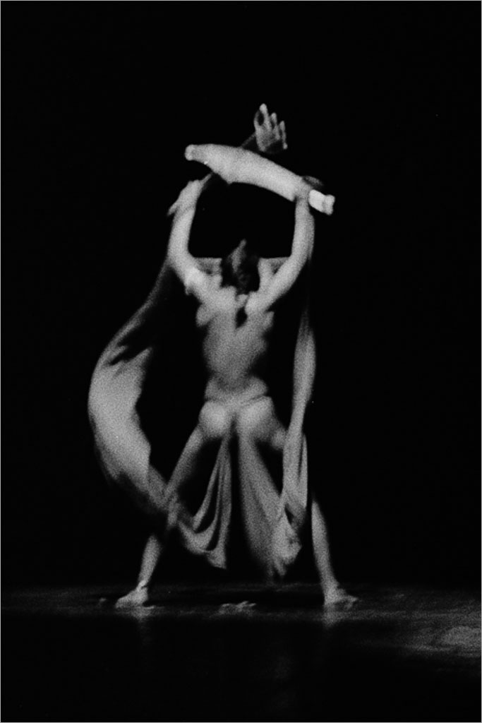 Uma imagem borrada de uma artista no palco, em meio a um movimento. Os braços da artista estão levantados, segurando um tecido fluido, criando um movimento dinâmico contra um fundo escuro. Foto: Guto Muniz