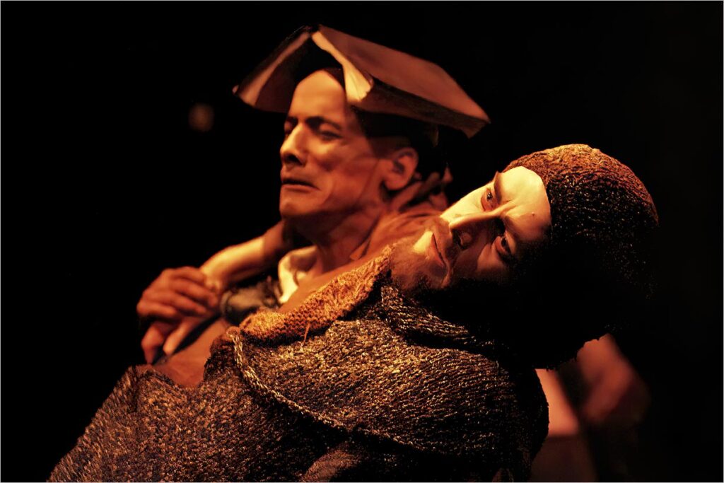 Dois atores no palco, um com um livro aberto na cabeça, ambos vestidos com trajes medievais. A cena é tem uma iluminação suave, criando uma atmosfera dramática. Um ator parece contemplativo enquanto o outro se inclina para frente com uma expressão intensa. Foto: Guto Muniz