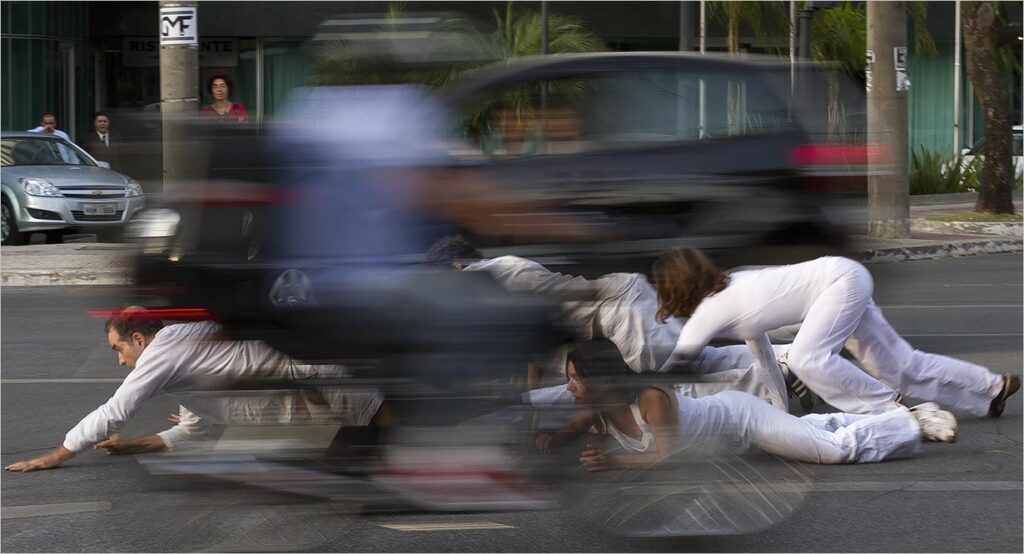 Um grupo de pessoas, mulheres e homens, vestindo branco rastejam em uma rua enquanto um motociclista e um carro borrados passam. O fundo mostra uma vitrine e árvores. A cena transmite movimento e uma performance de rua incomum. Foto: Guto Muniz