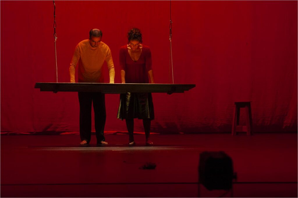 Um homem e uma mulher, estão de pé próximas um do outro no palco sob iluminação vermelha, cada um com as mãos apoiadas sob a mesa suspensa. Um banco é visível ao fundo. Foto: Guto Muniz
