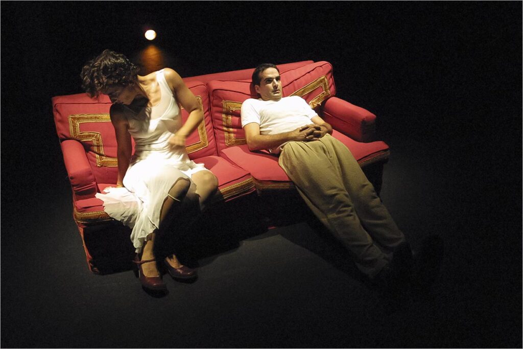 Uma mulher de vestido branco está sentada em um sofá vermelho, olhando para baixo, enquanto um homem de camisa branca e calça bege está deitado na extremidade oposta, olhando para cima. A cena é pouco iluminada e tem uma fonte de luz atrás deles. Foto: Guto Muniz