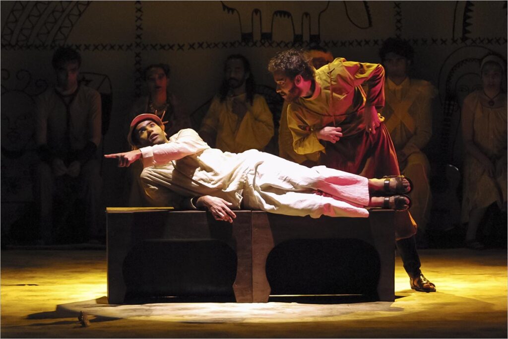 Uma cena teatral de palco apresentando um homem em vestes brancas deitado sobre uma mesa, apontando para a esquerda. Outro homem em um traje marrom e amarelo se inclina sobre ele com uma expressão intensa e curiosa. Várias pessoas sentam-se no fundo pouco iluminado contra uma parede decorada na penumbra. Foto: Guto Muniz