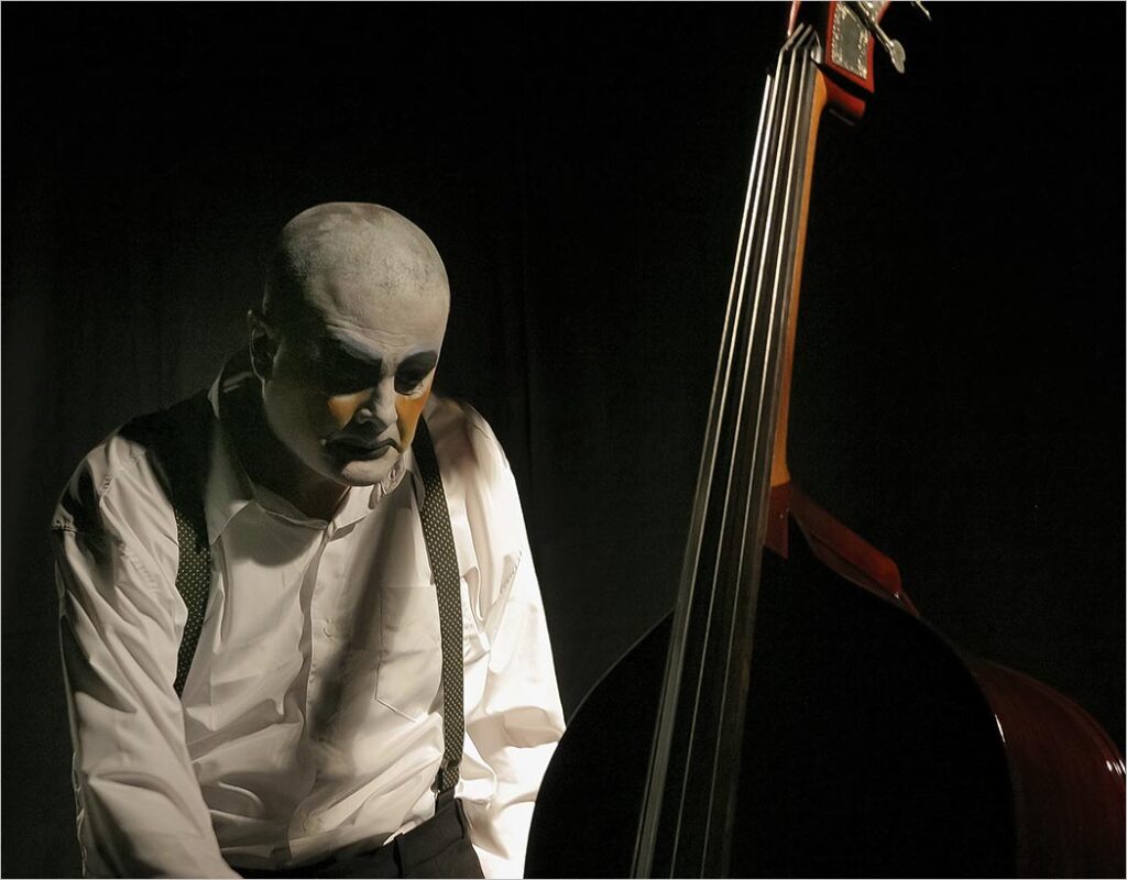 Um homem com maquiagem de palhaço preta e branca, vestindo uma camisa branca e suspensórios, está sentado em uma sala sutilmente iluminada, olhando para baixo, ao lado de um grande contrabaixo. A iluminação projeta sombras dramáticas contra um fundo preto. Foto: Guto Muniz