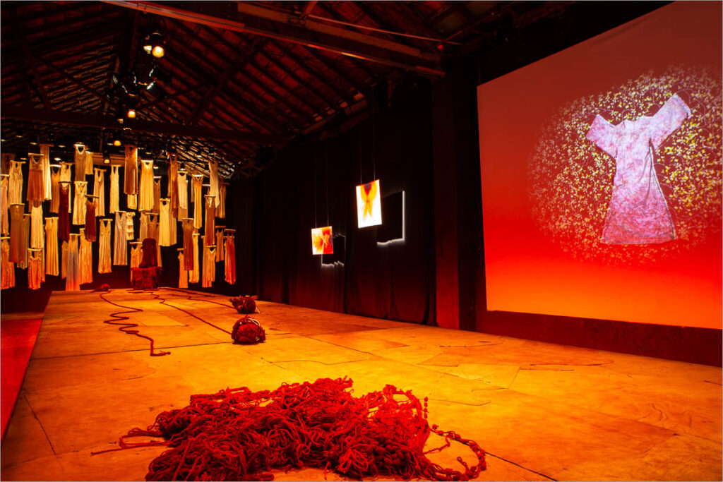 Uma sala iluminada por uma luz alaranjada e dispersa apresenta um palco, como uma passarela, com vestidos suspensos pelos cabides ao fundo e iluminação avermelhada. Uma tela grande, na lateral do palco, exibe a imagem de um vestido. Pilhas de um fio vermelho repousam no chão de maneira desorganizada. Foto: Renato Mangolin
