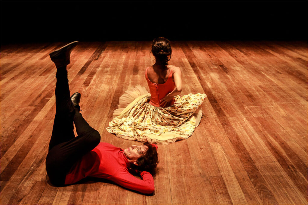 Uma mulher mais velha, de camisa vermelha de manga longa e calça preta, está deitada em um palco de madeira com as pernas levantadas ao alto e aos mãos atrás de sua cabeça como um travesseiro. Uma de suas pernas se encontra dobrada com o tornozelo sobre o joelho da perna esticada para cima. Seu corpo está virado para a esquerda da imagem. De costas para a câmera, um pouco mais ao fundo, uma outra mulher, usando um vestido de saia estampada de flores e corpete vermelho, se encontra sentada no chão, com postura ereta, cabelo preso e uma de suas mãos em suas costas. O palco possui uma iluminação quente e dispersa com o fundo escuro. Foto: Renato Mangolin