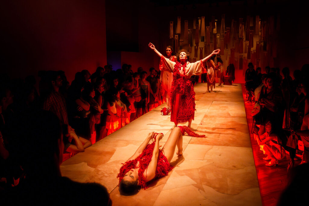 Artistas em trajes vermelhos com branco, texturizados, atuam em uma passarela cercados por uma plateia. Uma mulher fica de pé com os braços levantados, e outra deita no chão a sua frente. A cena é iluminada por luzes vermelhas e quentes. Ao fundo se vê vários vestidos suspensos pelos cabides de forma dispersa enquanto na penumbra juntamente com outras figuras. Foto: Renato Mangolin