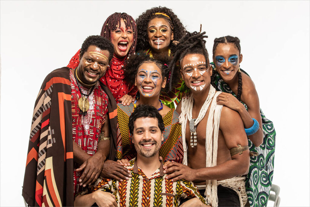 Um grupo diverso de sete pessoas vestindo roupas vibrantes e tradicionais africanas posa junto, sorrindo. Eles têm pintura facial colorida e estão posicionados contra um fundo branco simples. Foto: Renato Mangolin