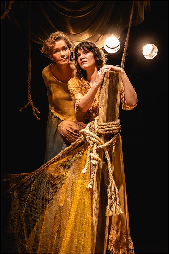 Duas mulheres estão em uma cena iluminada com luz quente e focada, cercadas por cordas e redes no que ilustra a proa de um navio. Uma se apoia em um toco de madeira, enquanto a outra está atrás dela. Elas usam vestidos fluidos em tons terrosos e são iluminadas por luzes no teto. Foto: Renato Mangolin