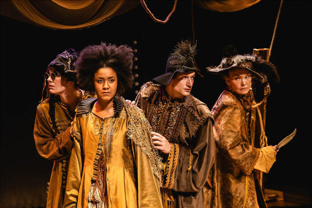 Quatro atores, homens e mulheres, em trajes de época ornamentados, marrom-dourados, atuam no palco contra um fundo escuro. Uma atriz olha para frente, enquanto os outros olham em direções diferentes com expressões dramáticas, iluminados suavemente por uma luz quente. Foto: Renato Mangolin