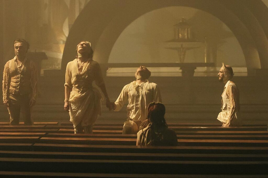 Quatro pessoas em trajes teatrais estão dramaticamente de pé com os braços ao lado do corpo entre bancos de igreja, de frente para direções diferentes. Outra pessoa está sentada de costas para o espectador, olhando para elas. O cenário tem uma iluminação atmosférica e dourada. Foto: Guto Muniz