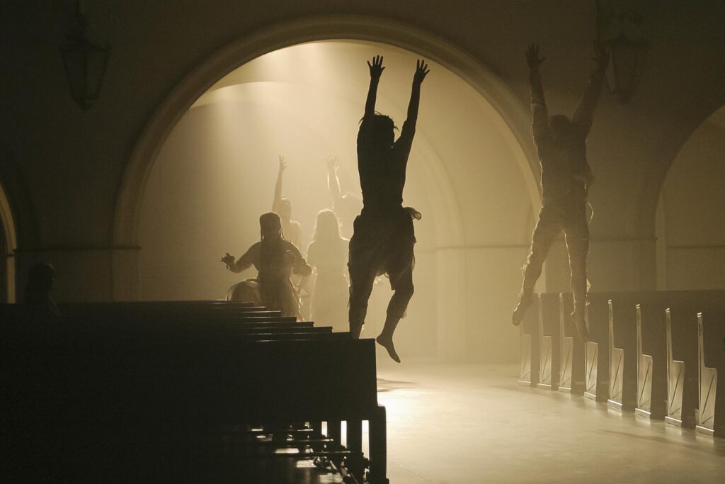 Atores em silhueta saltam em um salão de iluminação baixa, com portas em arco e luz dispersa, criando uma atmosfera dramática. Duas figuras em primeiro plano estão no ar, braços esticados para cima, enquanto outras se movem no fundo. Foto: Guto Muniz