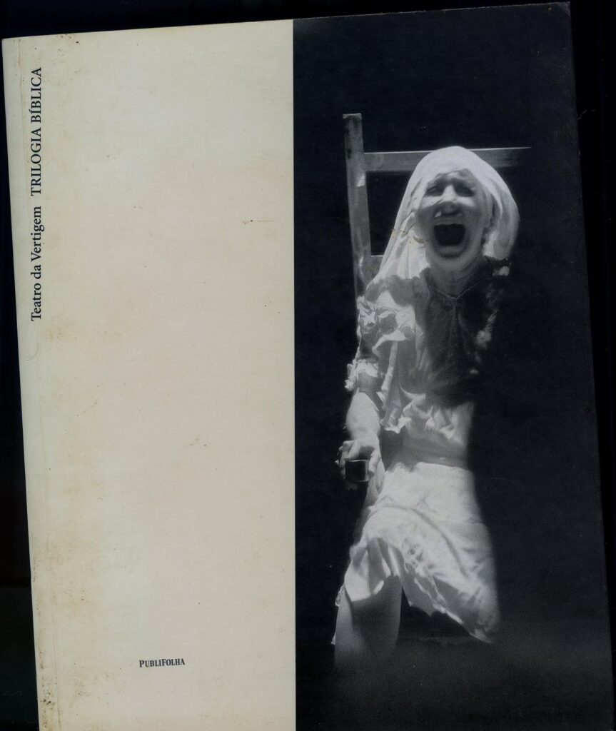 Uma imagem em preto e branco captura uma pessoa com uma expressão aflita e boca aberta, possivelmente gritando ou berrando. Vestida de branco com um lenço na cabeça, ela está parcialmente na sombra. O texto à esquerda diz Teatro da Vertigem TRILOGIA BÍBLICA, destacando um espetáculo do Teatro da Vertigem.