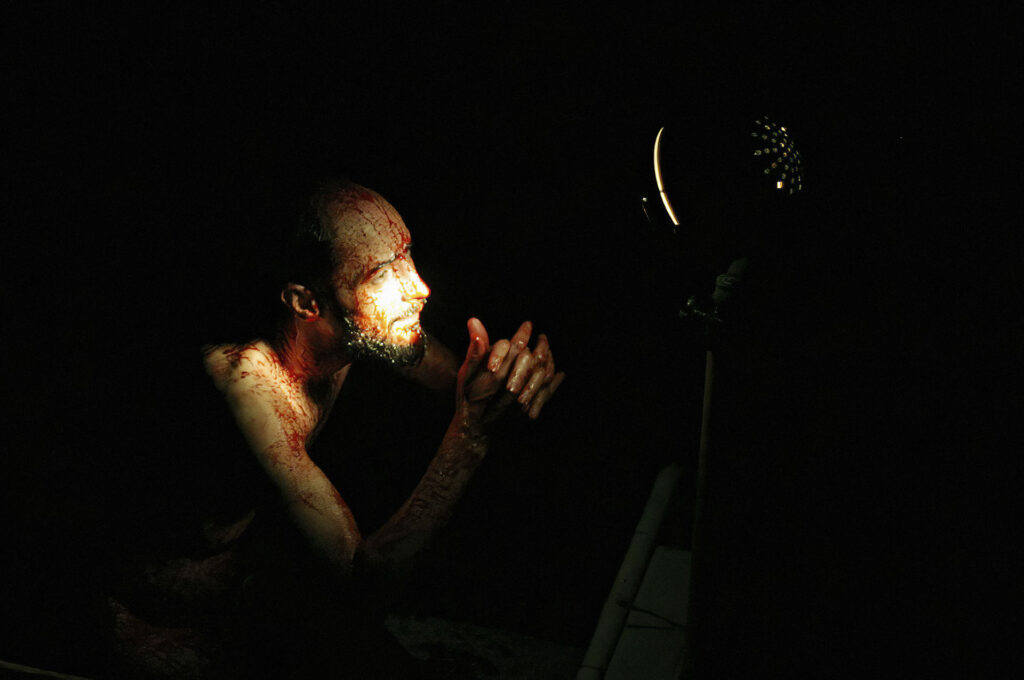 Uma pessoa com o rosto sujo de sangue é iluminada por uma única fonte de luz em um ambiente escuro. Ela está juntando as mãos e olhando em direção à luz, projetando sombras em suas feições. O fundo está quase todo obscurecido pela escuridão. Foto: Guto Muniz