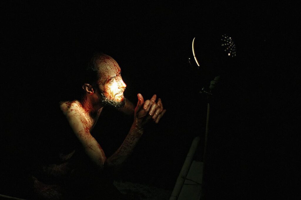 Uma pessoa com o rosto sujo de sangue é iluminada por uma única fonte de luz em um ambiente escuro. Ela está juntando as mãos e olhando em direção à luz, projetando sombras em suas feições. O fundo está quase todo obscurecido pela escuridão. Foto: Guto Muniz