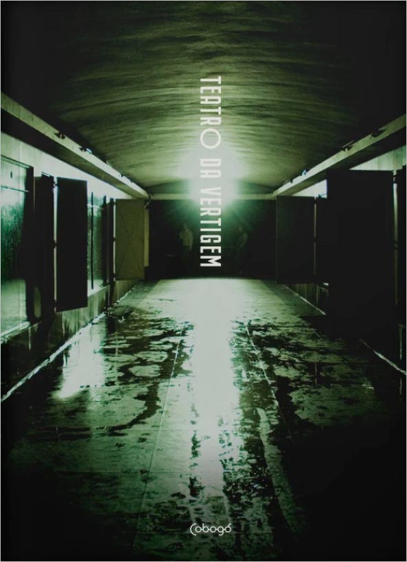 Um corredor estreito e pouco iluminado com água refletida no chão e iluminado por uma luz esverdeada brilhante no final. O texto vertical diz Teatro da Vertigem.