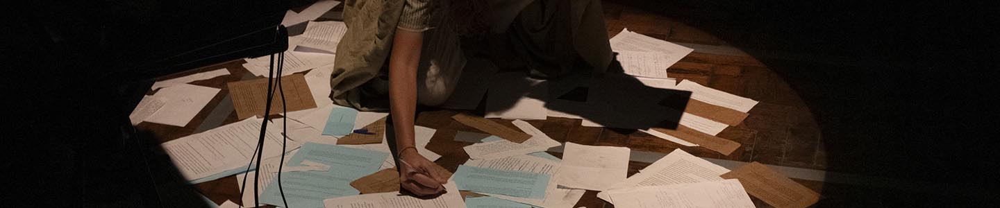 Uma mulher se ajoelha sob um holofote em um palco pouco iluminado, cercada por papéis espalhados. Ela está segurando alguns papéis e escreve eu um que está no chão. Foto: Jorge Alves