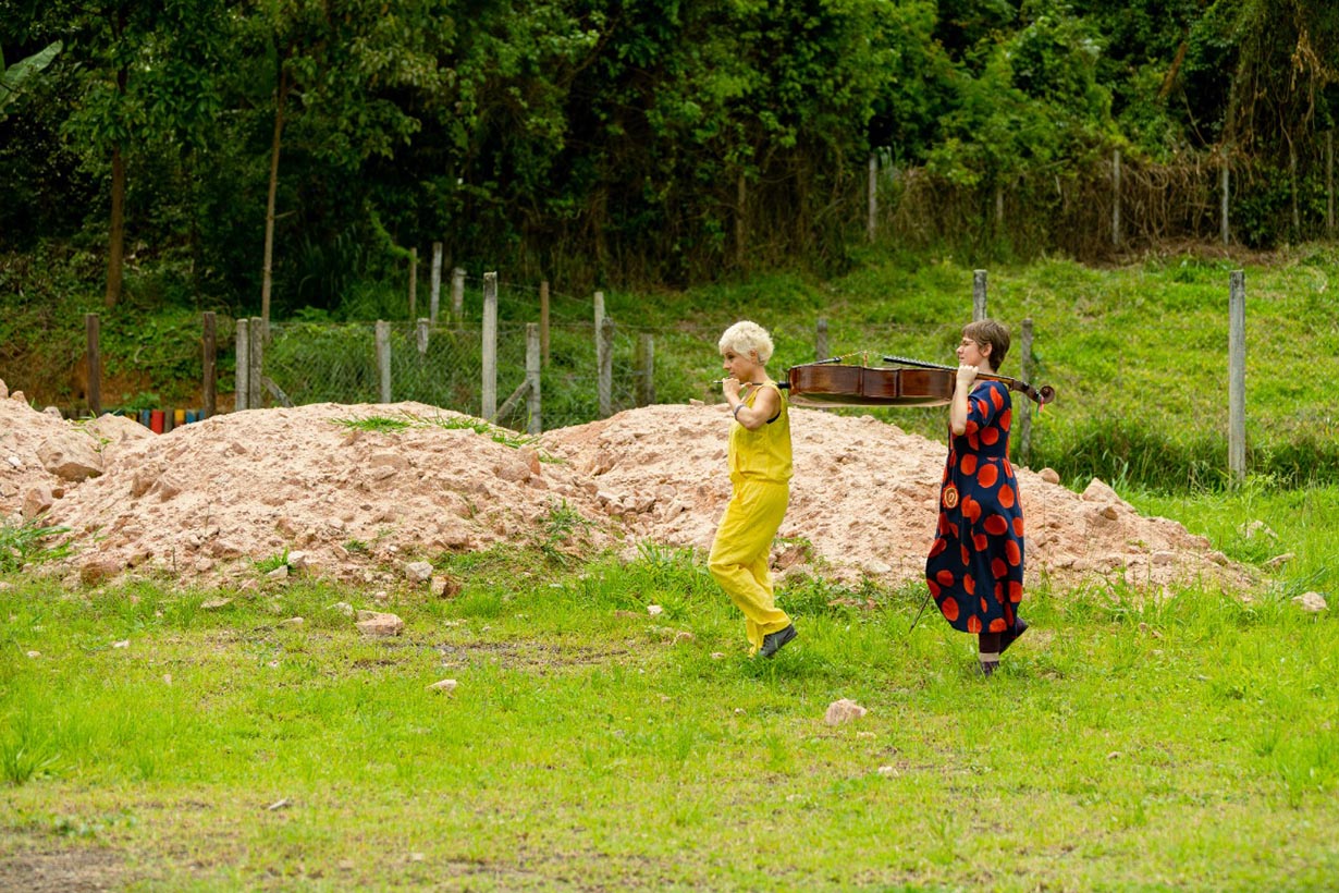 Duas mulheres caminhando na grama perto de montes de areia. Uma usa um macacão amarelo e a outra usa um vestido escuro com círculos vermelhos. Elas seguram um mesmo violoncelo nos ombros. Árvores e uma cerca estão ao fundo. Foto: Thaís Grechi
