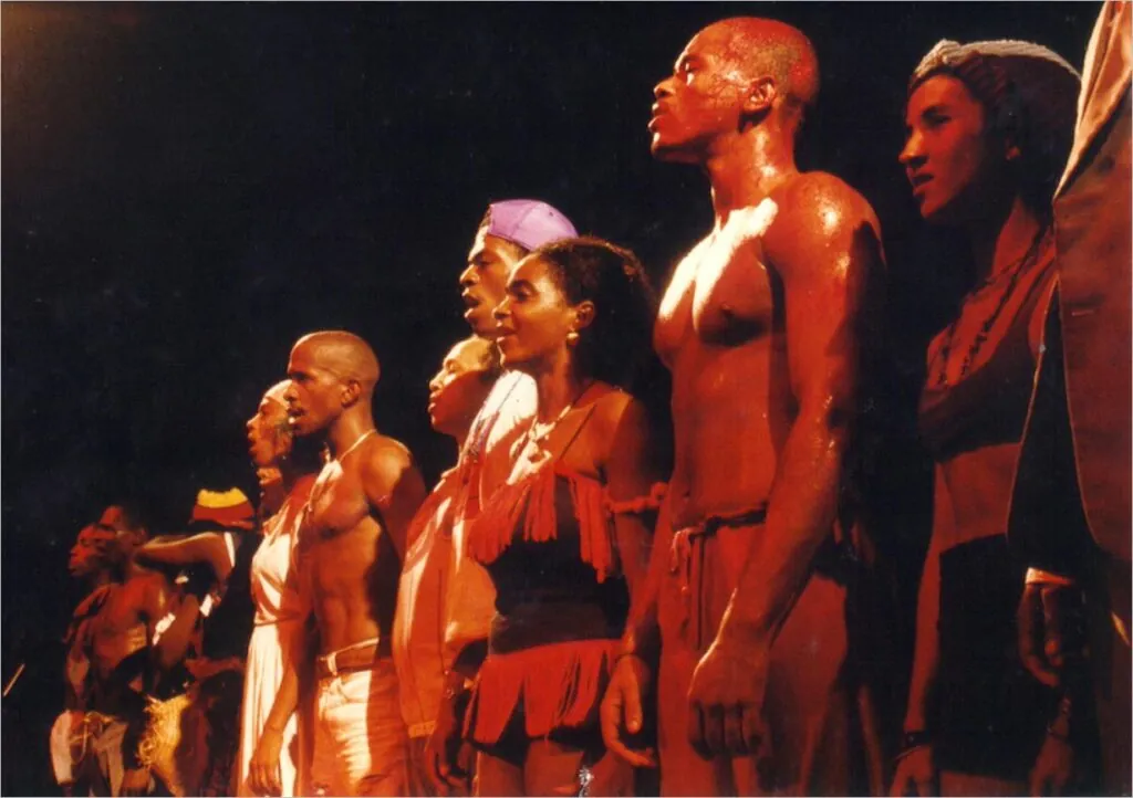 Um grupo de artistas fica lado a lado no palco, voltado para a frente. Eles usam trajes e acessórios de cabeça coloridos e minimalistas, com uma iluminação dramática que destaca suas expressões e corpos. Foto: Isabel Gouveia