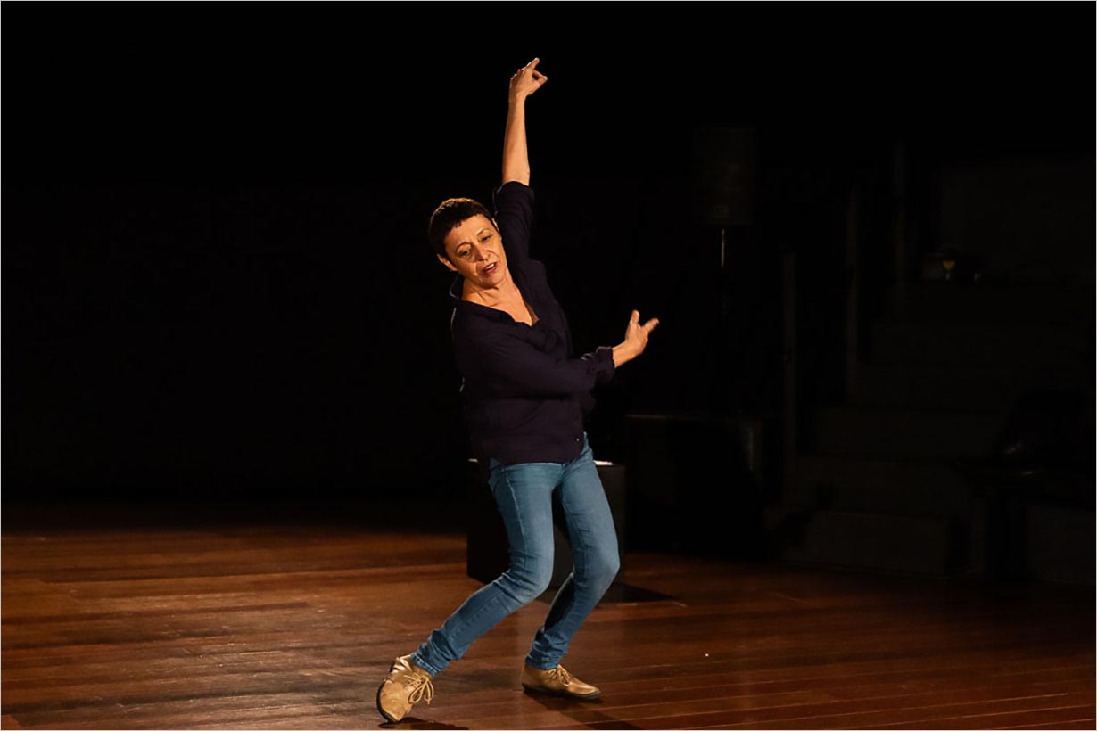 Uma mulher em um palco de madeira faz uma pose de dança com um braço levantado e o outro dobrado, vestindo uma camisa escura, calça jeans azul e sapatos bege, com um fundo escuro atrás dela.