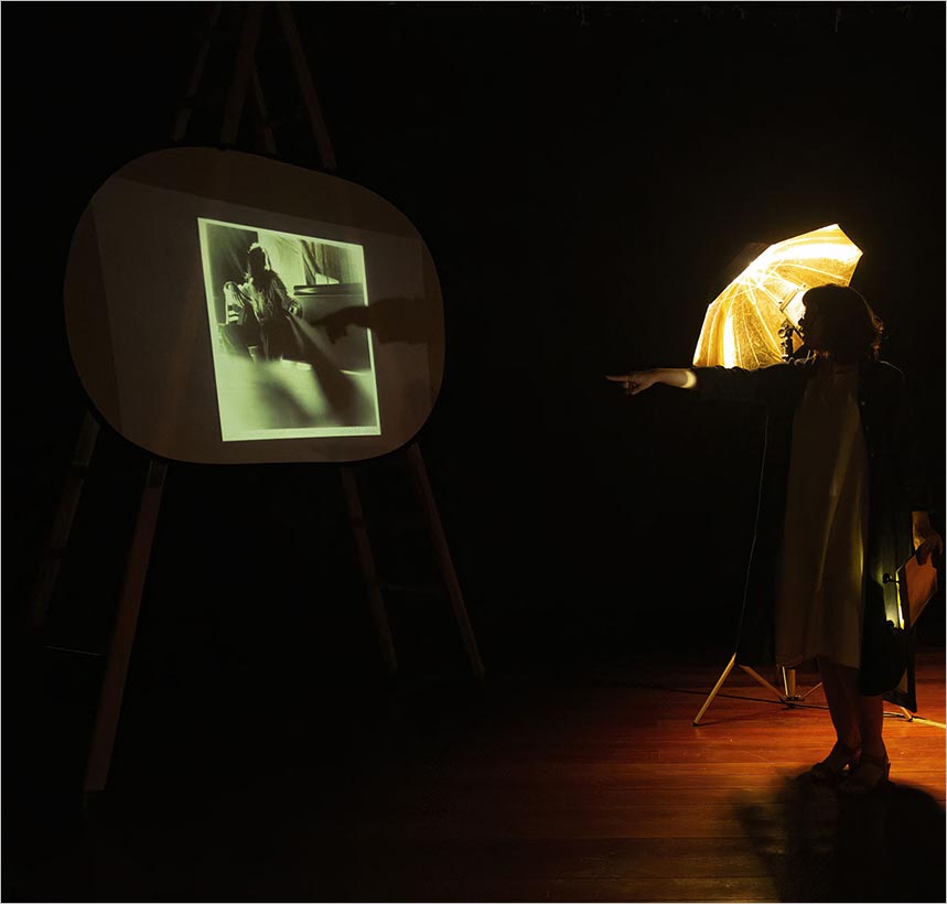 Uma pessoa está próxima a uma sombrinha dourada iluminada, apontando para uma foto em preto e branco projetada em uma tela em uma sala pouco iluminada com piso de madeira. Foto: Guto Muniz
