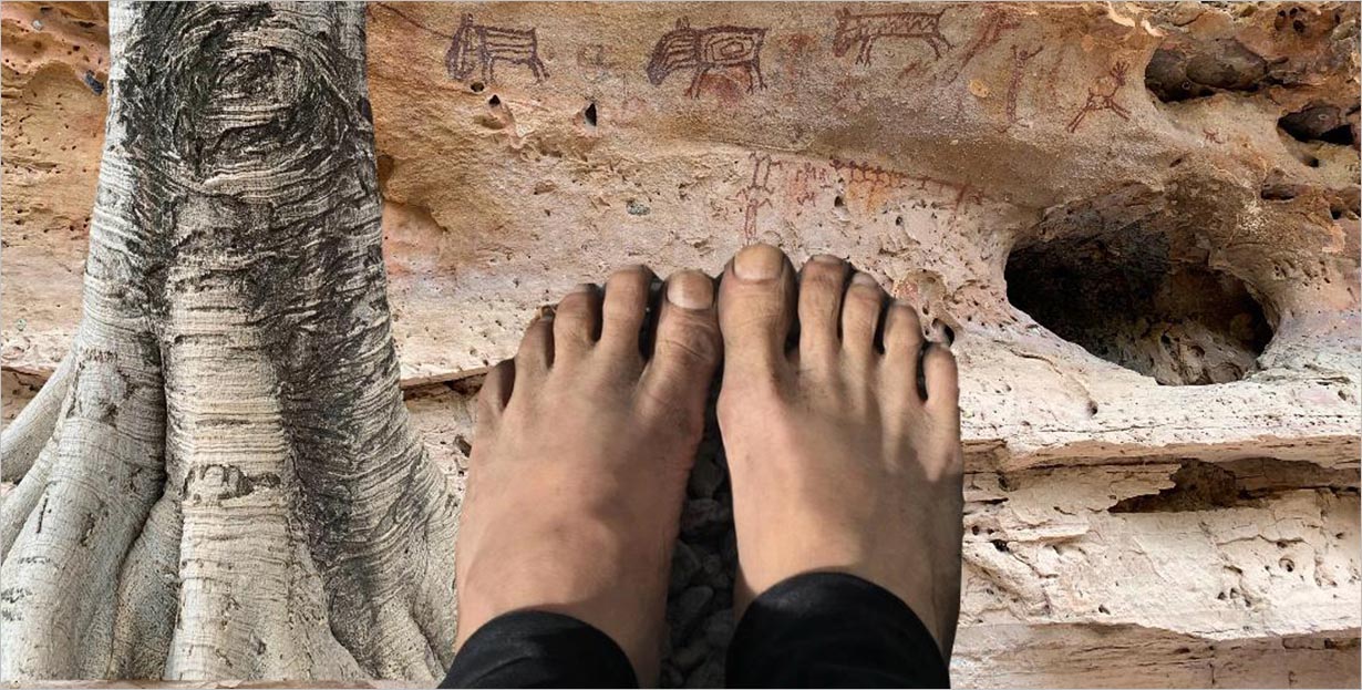 Pés descalços são mostrados em pé sobre um solo rochoso em frente a uma parede de rocha desgastada pelo tempo com esculturas antigas ou pinturas rupestres e um close-up de um tronco de árvore no lado esquerdo da imagem.
