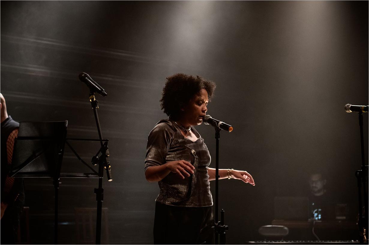Uma mulher canta em um microfone em um palco. Dois outros microfones e um suporte para partituras estão próximos, e uma pessoa trabalha em um laptop ao fundo. Foto Íra Barillo
